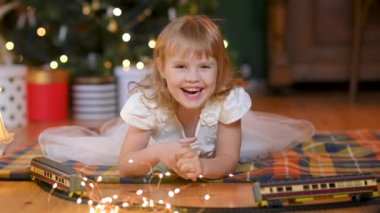 Şenlikli bir Noel ağacının arka planında, yerde, elbiseli, sevimli, mutlu bir kız gülümseyip kameraya bakıyor. Çocuk konseptiyle yeni yıl ve tatiller.