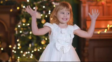 Beyaz elbiseli şirin küçük bir kız Noel ağacının arka planında oynuyor. Çocuk kar taneleri gibi düşen beyaz tüyleri yakalıyor. Pozitif duygular.