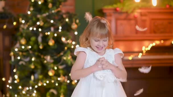 白いドレスを着た可愛い女の子がクリスマスツリーを背景に部屋で遊んでいます 子供は雪のフレークのような白い羽をキャッチします 肯定的な感情 — ストック動画