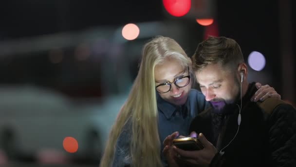 若いカップルは夜の街の通りに立っている間にスマートフォンを使用します 画面からの光 通信オンライン 技術コンセプト — ストック動画