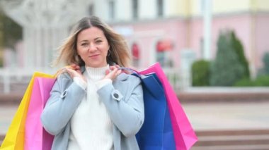 Şehirde alışveriş torbalarıyla dolaşan ceketli çekici bir kızın portresi. Alışveriş meraklısı genç bir kadın, elinde alışveriş torbalarıyla kameraya gülümsüyor.