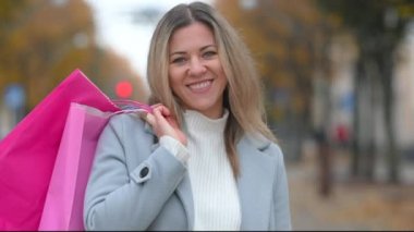 Genç, mutlu, alışveriş düşkünü, caddede yürüyen ve akıllı telefonda yazan bir kadın. Gülümseyen, sohbet eden ve alışveriş torbaları tutan bir bayan..