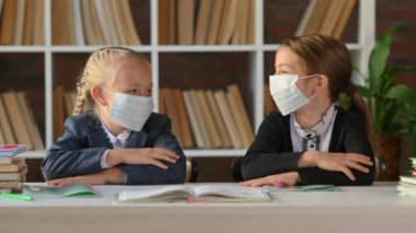 Koruyucu tıp maskeli genç okul kızları sınıftaki masa başında oturup gülüyorlar. çocuk eğitim kavramı