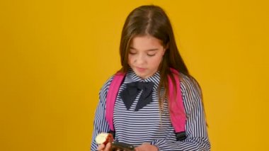 Sırt çantalı bir kızın portresi elma yiyor ve sosyal ağlarda iletişim kuruyor. Şirin kız telefonda sarı arka planda mesaj yazıyor. teknoloji ve iletişim konsepti