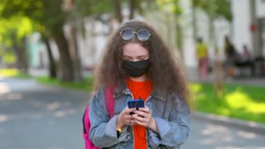 Şehir Sokağında Akıllı Telefonla İletişim kuran Koruyucu Maskeli Genç Çekici Kız. Z jenerasyonu şehirdeki telefonla etkileşime geçiyor. Telefondaki Kadın Mesajları Açık havada.