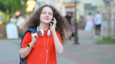 Genç Çekici Kız Smartphone 'da Sokakta Yürüyen Şehir Haberleşmesi. Boynunda kulaklık olan kız cep telefonu kullanıyor, konuşuyor ve gülümsüyor..