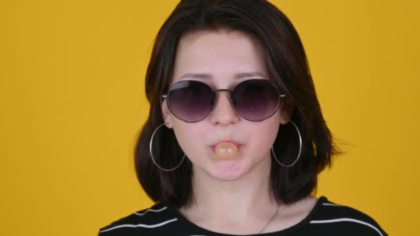 サングラスガムを噛むとスタジオの黄色の背景に泡を吹いて10代の女の子の肖像画 人々のライフスタイルコンセプト — ストック動画