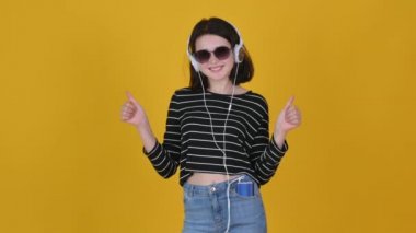 Güneş gözlüklü genç bir kızın portresi kulaklık takıyor müzik dinliyor ama akıllı telefon gülümsüyor ve baş parmak işareti gösteriyor. Stüdyo fotoğrafçılığı sarı arka planda izole edilmiş. yaşam tarzı konsepti