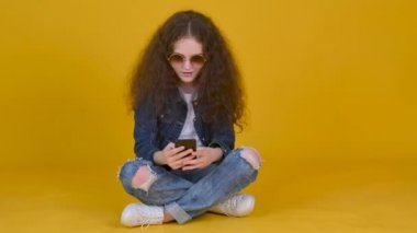 Kıvırcık saçlı, gözlük takan genç bir kız, sarı arka planda izole bir şekilde yerde otururken uygulamalarla iletişim kuran akıllı telefonlar kullanıyor..