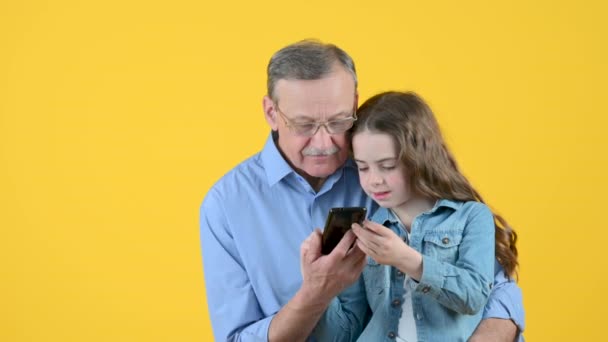 幸せな祖父とかわいい孫娘は黄色の背景に隔離されたスマートフォンを使用しています 孫娘は祖父にスマートフォンを使うように教えています — ストック動画