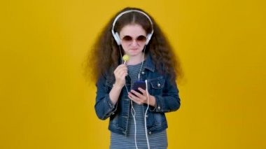 Beyaz kulaklıklı, şeker ve akıllı telefonlu, kıvırcık saçlı genç kız sarı bir stüdyoda müzik dinliyor..