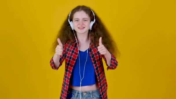 ヘッドフォンを身に着けている巻き毛を持つ若い面白い女の子は黄色の背景スタジオに隔離された親指のサインアップを示すカメラの外観 親指を出すカメラを見て — ストック動画