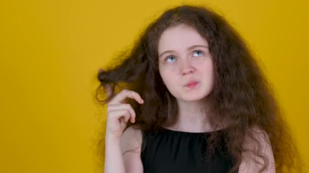 巻き毛とドレスを着た若い女の子は 黄色いスタジオの背景に隔離された 集中的な表情で脇を見て彼女の指で彼女の髪を回します 頭の良いアイデアを — ストック動画