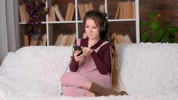 Красивая Молодая Женщина Дредами Слушает Музыку Черными Наушниками Сидя Диване — стоковое видео