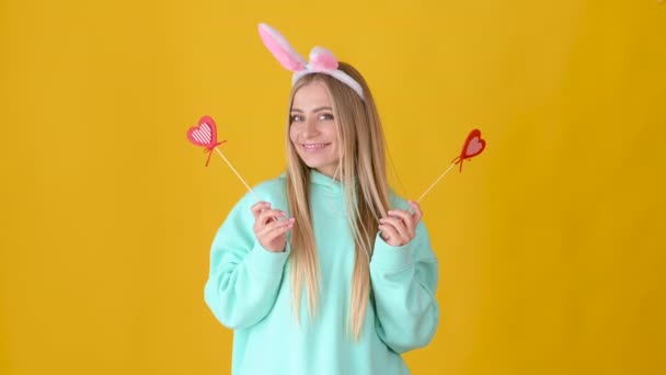 一个快乐的金发碧眼的小女孩的画像 她长着粉红的兔子耳朵 手里拿着一颗红心 在黄色的背景下孤独地笑着 情人节的概念 母亲节 复活节 — 图库视频影像