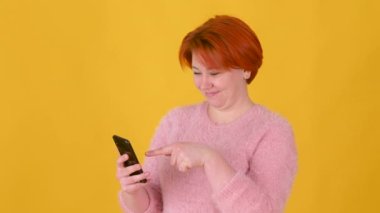 Parlak saçlı, akıllı telefon kullanan ve internette mesajlaşan mutlu genç kız sarı arka planda izole edilmiş bir gülümsemeyle. Halk yaşam tarzı konsepti.