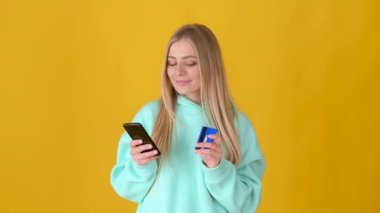 Cep telefonlu kredi kartı kullanan mutlu genç sarışın kız sarı arka planda online alışveriş yapıyor. Halk yaşam tarzı konsepti.