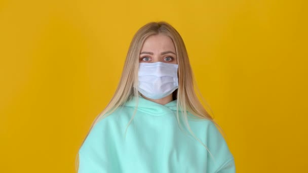 黄色のスタジオの背景に隔離された医療マスクの若いブロンドの女の子の肖像画 社会的距離と新常態 — ストック動画
