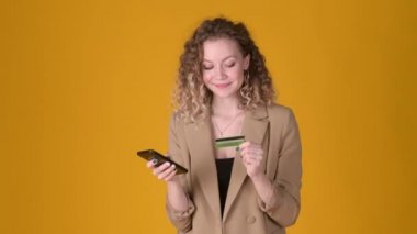 Kıvırcık saçlı neşeli ve cep telefonu kullanan genç bir kız kredi kartı tutuyor ve sarı arka planda internetten alışveriş yapıyor. Halk yaşam tarzı konsepti.