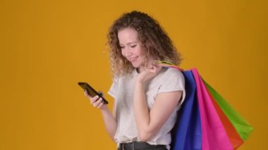 Alışveriş torbaları tutan kıvırcık saçlı genç kız sarı arka planda izole edilmiş arkadaşlarıyla akıllı telefon iletişimi kullanıyor. alışveriş kavramı