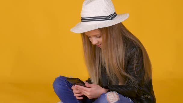 Маленькая Блондинка Шляпе Используя Мобильный Телефон Сидящий Жёлтом Фоне Концепция — стоковое видео