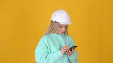 Genç pozitif düşünen miğferli kız, sarı arka planda izole edilmiş akıllı telefon düşünceleri kullanıyor..