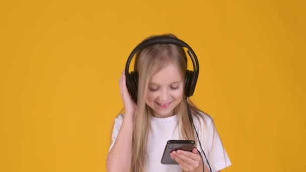 スマートフォンを持っている面白い小さなブロンドの女の子は ヘッドフォンで音楽に耳を傾け スタジオの黄色の背景で踊ります Mp3プレーヤーアプリケーション — ストック動画