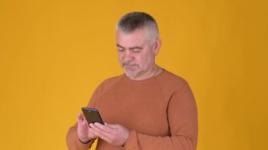 Yakışıklı yetişkin adam sarı stüdyo arka planında sosyal medyada akıllı telefon mesajları kullanıyor. Yaşlılar ve modern teknolojiler.