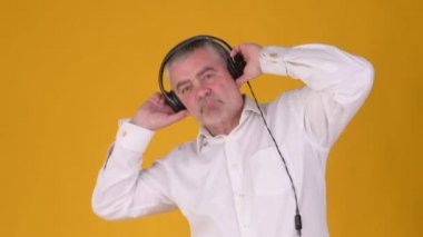 Beyaz tişörtlü, kulaklıklı, dans eden ve sarı arka planda izole edilmiş müziğin keyfini çıkaran mutlu, yaşlı, gri saçlı bir adam. Halk yaşam tarzı konsepti.
