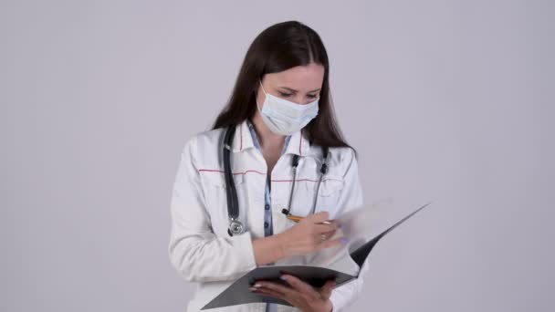 头戴外科口罩 头戴医用制服 有自信的女医生的画像 用白色背景分离出临床笔记 医务工作者 — 图库视频影像