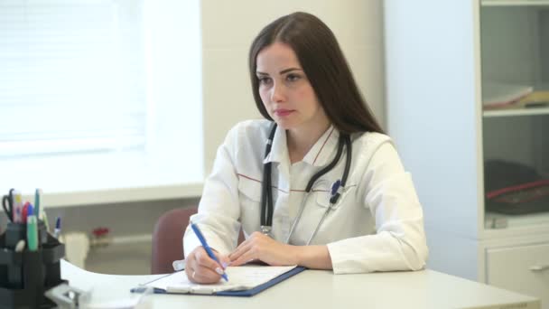 医学の女性医師制服は 患者に耳を傾け お勧めします 若いセラピストがデスクに座っている医療チェックリストを記入しています 健康と安全の概念 — ストック動画