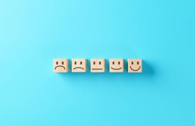 Müşteri memnuniyeti ve ahşap bloklar ile geri bildirim kavramı mavi arka planda mutsuzdan mutluluğa farklı emoji yüzleri gösteren, kullanıcı deneyimini temsil eden ve derecelendirme ölçeğini gözden geçiren.