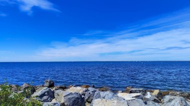 Bir gölün canlı mavi suyu ufukta gökyüzü ile buluşur, ön planda kayalık bir kıyı şeridi ve uzak suyu noktalayan küçük yelkenliler tarafından çerçevelenir.