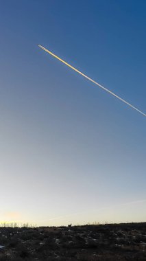 Uçağın izleri, batan güneşle aydınlanan açık mavi gökyüzü boyunca çapraz olarak çizilir. Aşağıdaki karanlık ufukta küçük bir geyiğin silueti görülüyor.