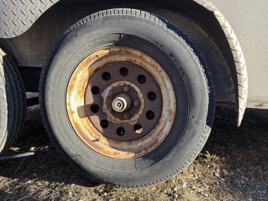 Ağır paslanmış metal kenarlı karavan lastiğinin yakın çekimi. Oksitlenme ve toprak ve çakıl yüzeyinde aşınma gösteren yıpranmış otomotiv ekipmanlarının detaylı bir çekimi.