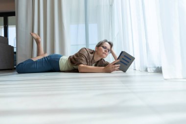 Evde kitap okuyan orta yaşlı bir kadın.