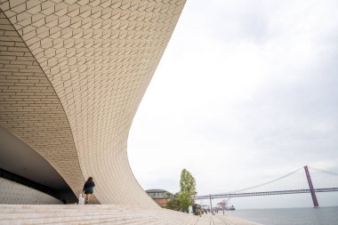 Tagus nehri boyunca uzanan 25 de Abril köprüsü Belem Lisbon Portekiz 'de bulutlu gökyüzünün altında MAAT Müzesi' nin kıvrımlı cephesi. 04 Kasım 2025.