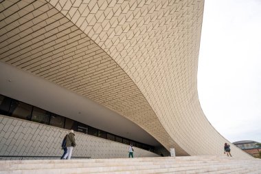 Belem Lisbon Portekiz 'de bulutlu bir günde MAAT Müzesi' nin eğimli seramik cephesinin önünde oturan ve dinlenen insanlar. 04 Kasım 2025.