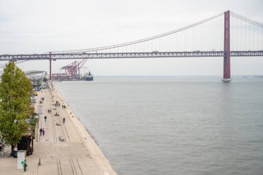Lizbon 'daki Belem rıhtımından görüldüğü gibi Tagus nehri üzerindeki 25 de Abril köprüsünün bulutlu gökyüzünün altında panoramik manzarası. 04 Kasım 2025.