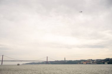 4 Kasım 2025 'te Lizbon' da bulutlu bir öğleden sonra gökyüzü altında Tagus Nehri üzerindeki 25 de Abril Köprüsü üzerinde uçan Kral anıtı İsa 'nın heykeli görülüyor..