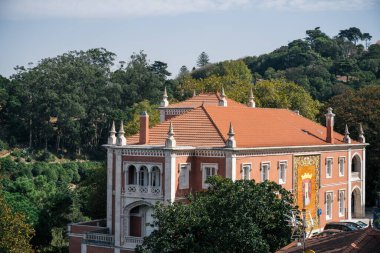 16 Ekim 2025 'te Sintra, Lizbon Bölgesi' nde yeşil orman tepelerine tepeden bakan beyaz kulesi olan renkli yamaç kenarlı evler..