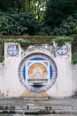 Portekiz 'in Sintra, Lizbon Bölgesi' nde 16 Ekim 2025 'te etrafı yeşilliklerle çevrili karmaşık mavi ve turuncu Portekiz fayansları ve yontulmuş meleklerle dolu taş çeşme..
