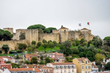 Lizbon Portekiz Aralık 05, 2025. Kale duvarları, Lizbon şehir merkezindeki tepedeki evler ve ağaçların üzerinde bulutlu gökyüzü koşulları altında yükseliyor..