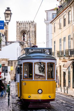Lizbon Portekiz Aralık 05, 2025. Sarı elektrikli tramvay, tarihi binaları ve taş kilise kulesi olan dar bir cadde boyunca ilerliyor..