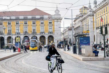 Lizbon Portekiz Aralık 05, 2025. Bir bisikletçi kalabalık bir şehir meydanında yayalar ve toplu taşıma araçlarıyla tramvay raylarından geçiyor..