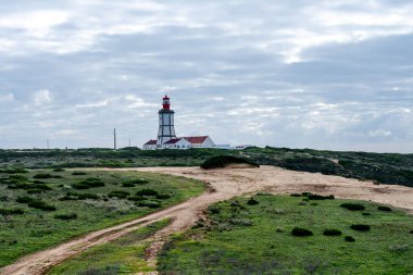 Sesimbra Portekiz 25 Aralık 2025. Bulutlu bir gökyüzünün altında Cabo Espichel deniz fenerine doğru uzanan alçak kıyı arazisinden geçen bir toprak yol var..