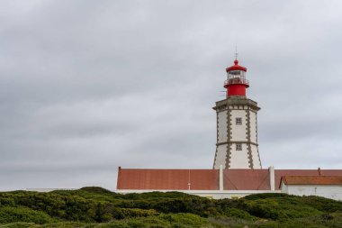 Sesimbra Portekiz 25 Aralık 2025. Cabo Espichel deniz feneri kulesi kıyı binalarının üzerinde bulutlu bir gökyüzüne karşı duruyor..