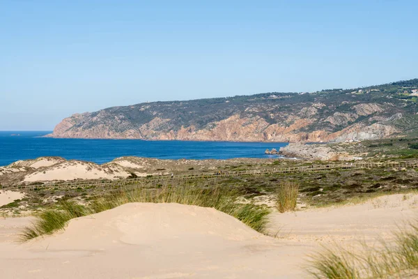 Sandy dune landscape with a wooden coastal path and distant rocky cliffs along the Atlantic coast near Guincho Cascais Portugal.
