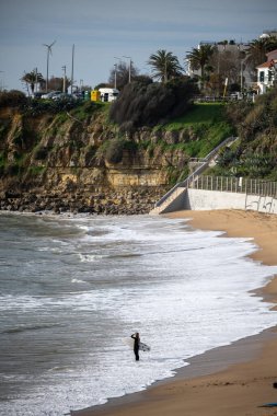 Estoril Lizbon Bölgesi, 17 Aralık 2025. Dalgalar kumlu kıyıya doğru yuvarlanırken bir sörf tahtası taşıyan bir sörfçü sığ sularda yürüyor..