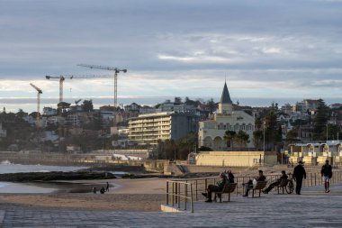 Estoril Lizbon Portekiz 17 Aralık 2025. İnsanlar arka planda iskan binaları ve inşaat vinçleri olan kumlu bir plajın yanındaki sahil boyunca oturur ve yürür..